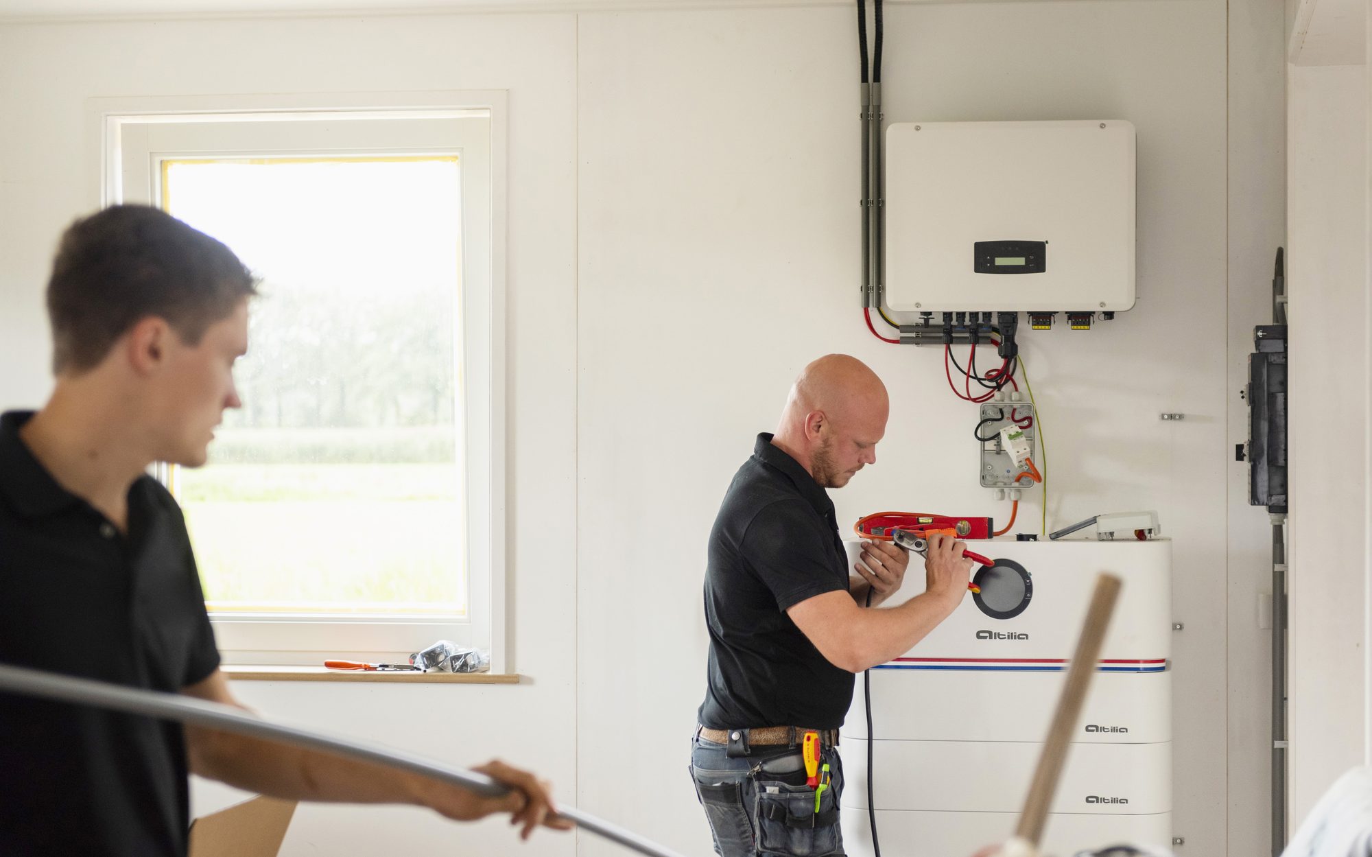 Twee installateurs aan het werk bij een Altilia-stapelbatterij, met de omvormer aan de wand erboven