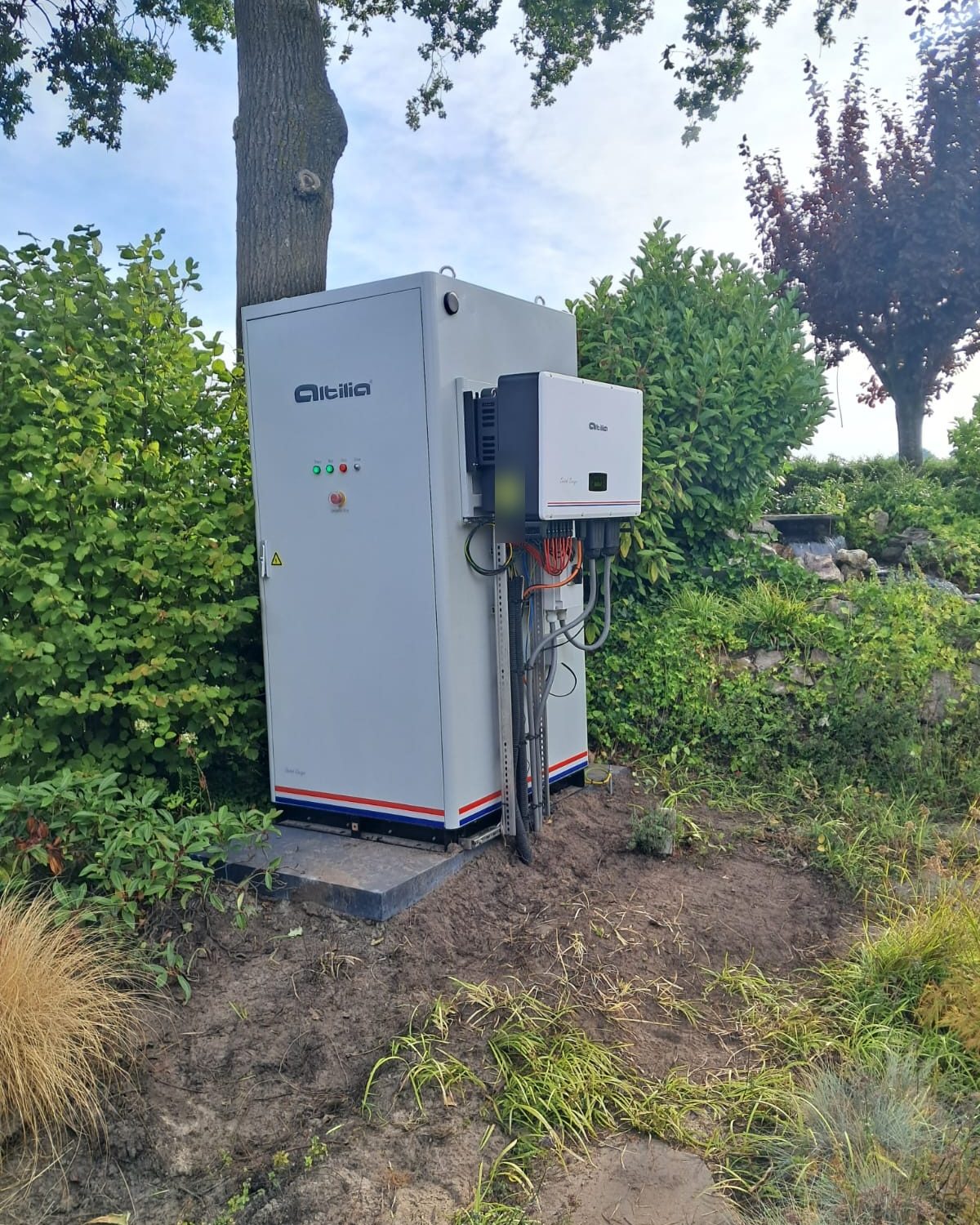 Altilia energiekast van 112 kWh buiten op een fundering tussen bomen en beplanting, met de omvormer aan de zijkant