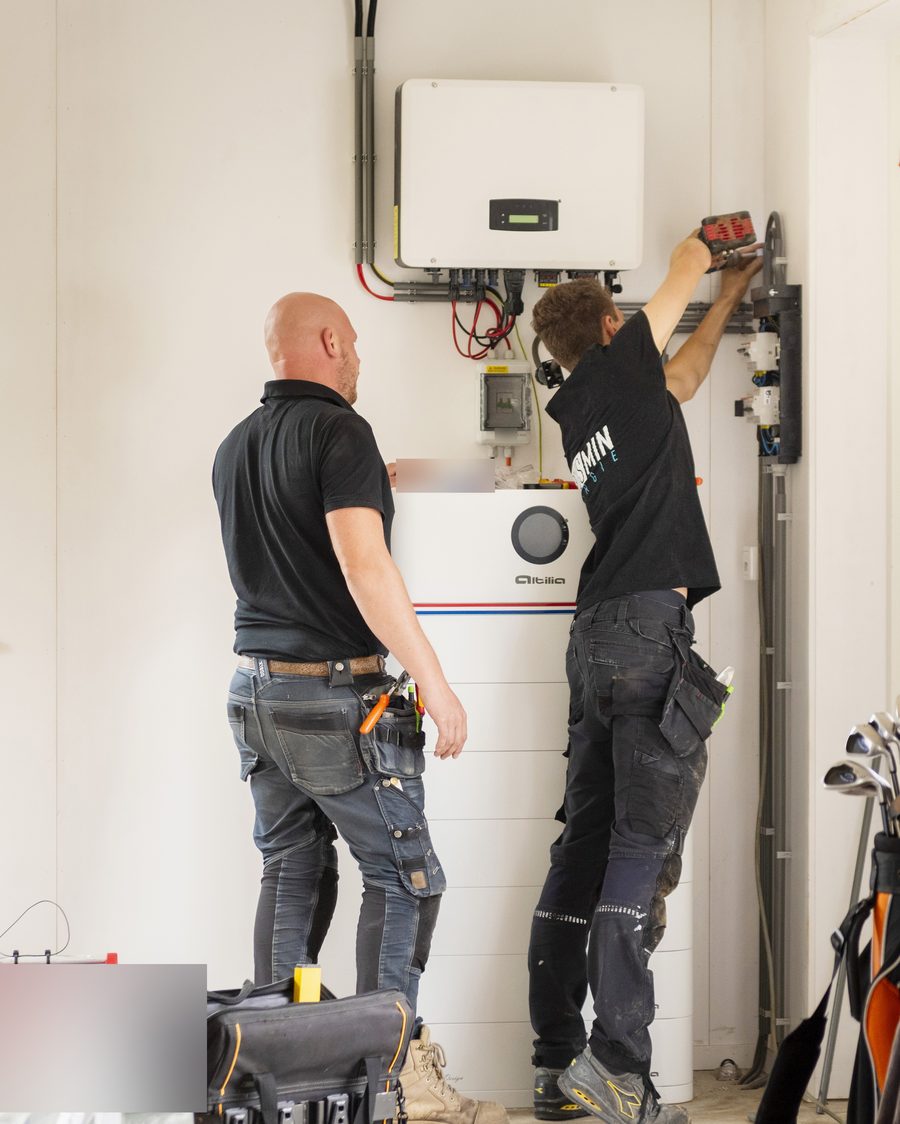Twee installateurs monteren een Altilia-thuisbatterij met de omvormer aan de wand van een berging
